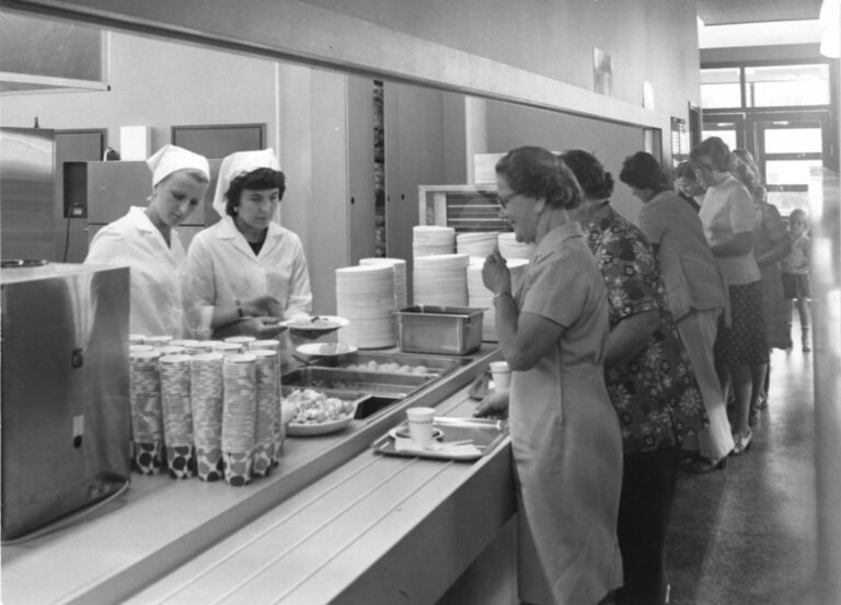 Ihmisiä jonottaa työpaikkaruokalan linjastossa tarjottimet edessään. Tiskin toisella puolella kaksi valkoisiin pukeutunutta työntekijää annostelee ruokaa lautasille. Vieressä monta pinoa lautasia sekä pahvikuppeja.