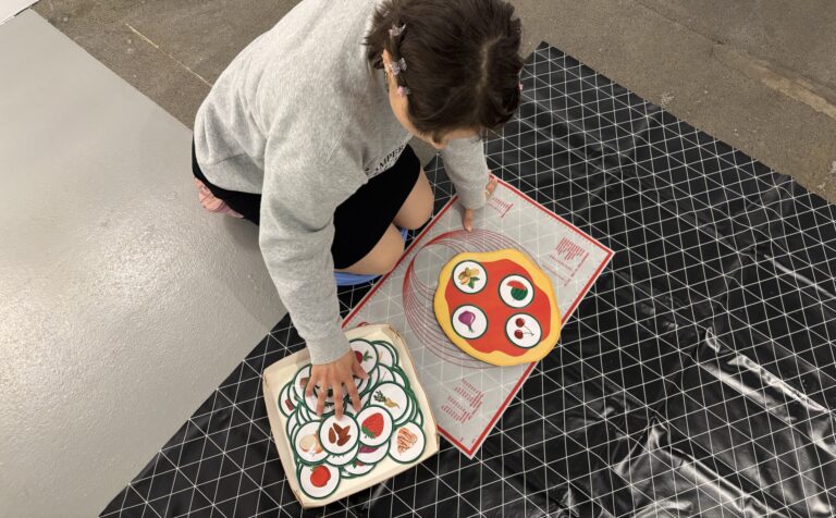 Henkilö kuvattuna ylhäältä päin asettelemassa kuvia erilaisista pizzatäytteistä paperisen pizzapohjan päälle.