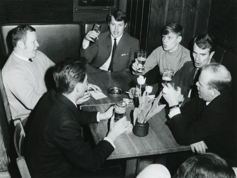 Ravintola-asiakkaita istuu pöydässä oluttuopit käsissään mahdollisesti laulaen.