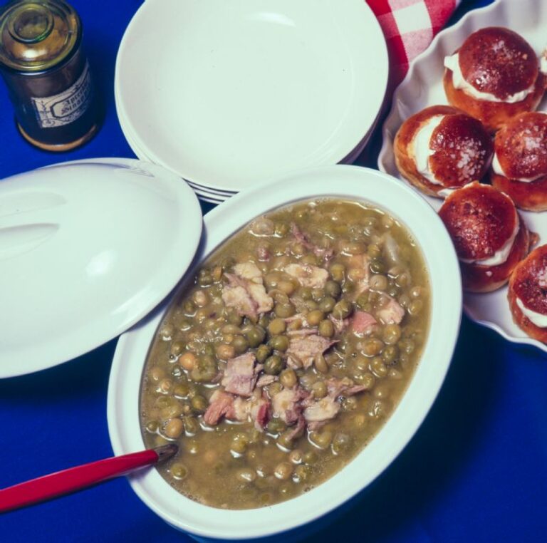 Valkoinen keraaminen pata, jossa hernekeittoa. Keitossa punavartinen kauha. Vieressä pino lautasia, padan kansi sekä vuoka, jossa laskiaispullia.