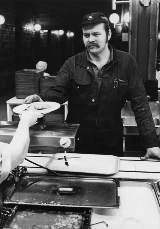 Haalariin ja lippalakkiin pukeutunut mies ottaa vastaan lautasen, jolla on ruokaa. Miehen edessä on tarjotin sekä vuokia, joissa on ruokaa.