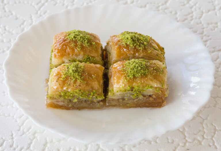 Valkoisella lautasella neljä pientä neliönmuotoista baklava-leivosta, joiden päällä vihreää pähkinörouhetta.