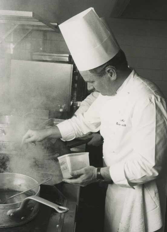 Valkoiseen kokin asuun ja hattuun pukeutunut mies pitää toisessa kädessä muovirasiaa ja ripottelee toisella kädellä jotakin isoon kattilaan. Taustalla näkyy keittiövälineitä ja -varusteita.