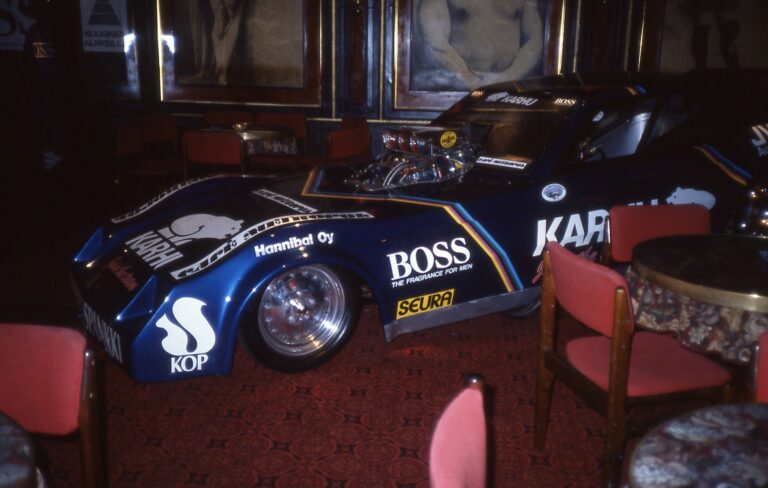 Ravintolasalissa oleva tummansininen urheiluauto, jossa Karhun, KOPin, BOSSin, Seuran ja Hannibal Oy:n logot. Ympärillä punaisella verhoiltuja tuoleja pyöreiden pöytien ympärillä. Taustalla näkyy seinällä isoja maalauksia.