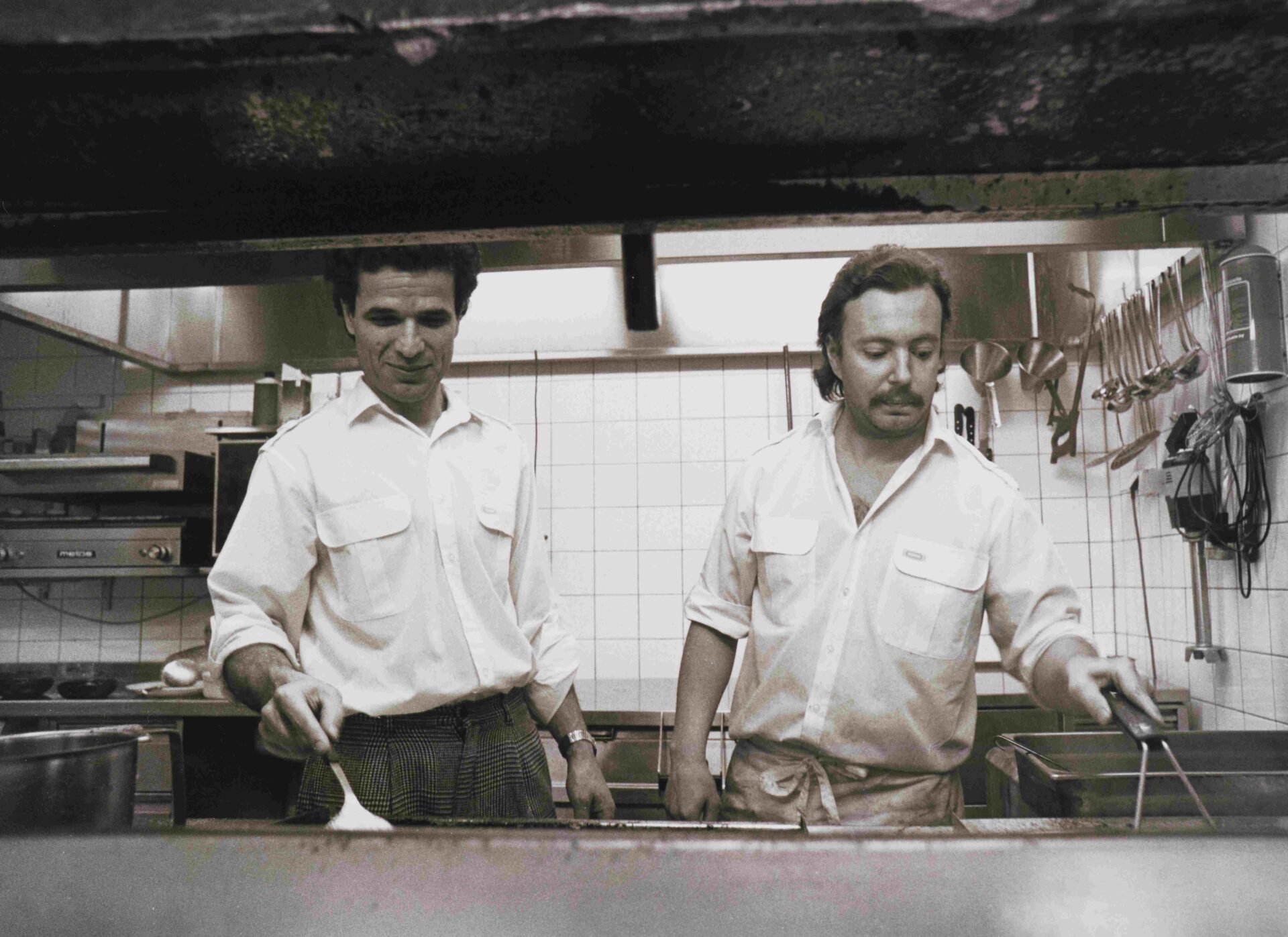 Kaksi miestä laittamassa ruokaa ravintolan keittiössä. Mustavalkoinen valokuva. Ravintola Österi Helsingin Itäkeskuksessa vuonna 1989.