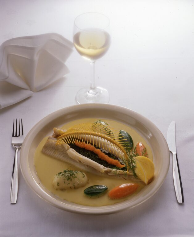 Lautanen, jolla kala-annos. Kalan ympärille levitetty kastiketta sekä erilaisia lisukkeita. Lautasen molemmin puolin aterimet ja edessä lasillinen valkoviiniä. Lautasesta vasemmalle yläviistoon valkoinen, viuhkaksi taitettu laustasliina.