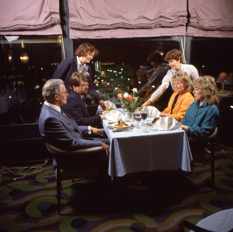 Kaksi naista ja kaksi miestä istuu ravintolapöydän ääressä. Pöydän taaimmaisen päädyn molemmin puolin kaksi naispuolista tarjoilijaa asettelee annoksia pöydälle asiakkaiden eteen. Pöydällä annoksia, lautasliinoja, viini- ja vesilaseja, ruokailuvälineet, tuhkakuppi ja kukkia maljakossa. Pöytä isojen ikkunoiden vieressä ja ikkunoissa liilat verhot. Taustalla näkyy öinen kaupunki.