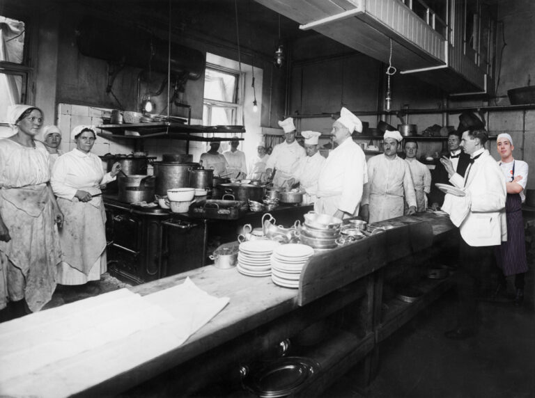 Kuvassa on vanha ravintolakeittiö ja sen henkilökuntaa 1920-luvulta. Historialliseen kuvaan on liitetty Ravintola Demon työskentelijä vuodelta 2015.