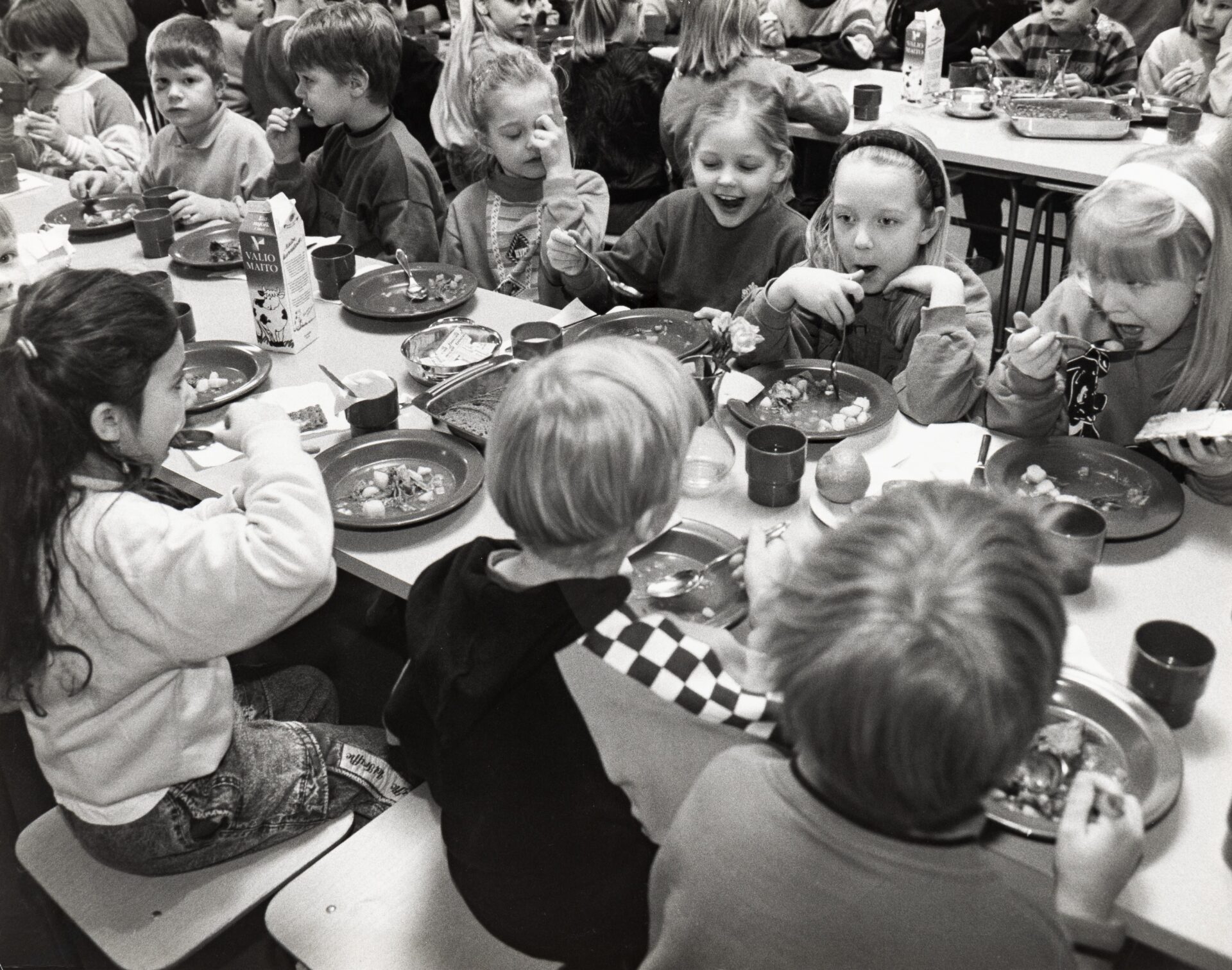 Lapsia istuu syömässä pitkän pöydän molemmin puolin. Pöydällä lautasia, joilla ruokaa, mukeja, hedelmiä, maitotölkki, leipää ja levitepakkauksia kulhossa. Taustalla näkyy toinen pitkä pöytä, jonka ääressä istuu lapsia syömässä.