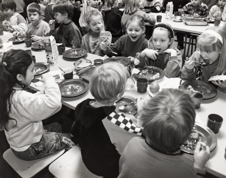 Lapsia istuu syömässä pitkän pöydän molemmin puolin. Pöydällä lautasia, joilla ruokaa, mukeja, hedelmiä, maitotölkki, leipää ja levitepakkauksia kulhossa. Taustalla näkyy toinen pitkä pöytä, jonka ääressä istuu lapsia syömässä.