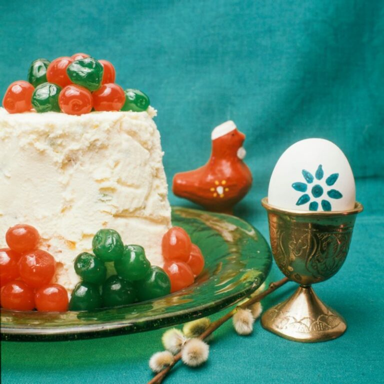 Vihreällä lasilautasella valkoinen pasha, joka koristeltu vihreillä ja punaisilla säilykekirsikoilla. Vieressä kananmuna metallisessa munakupissa, munaan maalattu vihreällä kukkakoristelu. Lisäksi pajunkissan oksa lautasen ja munakupin välissä sekä taustalla punainen kukkopilli. Vihreä kangas astioiden alla ja taustalla.