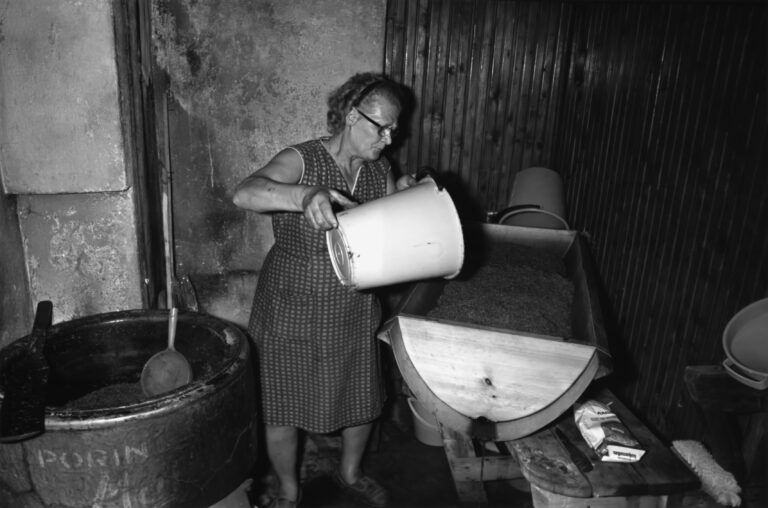 Nainen kaataa valkoisella ämpärillä vettä puiseen kuurnaan, jossa mäskiä. Kuurna makaa jonkinlaisella puisella penkillä. Vieressä muovisia astioita, puukko ja harja. Naisen toisella puolella iso pata, jossa iso kauha nojaa seinämään ja pohjalla mahdollisesti maltaita.