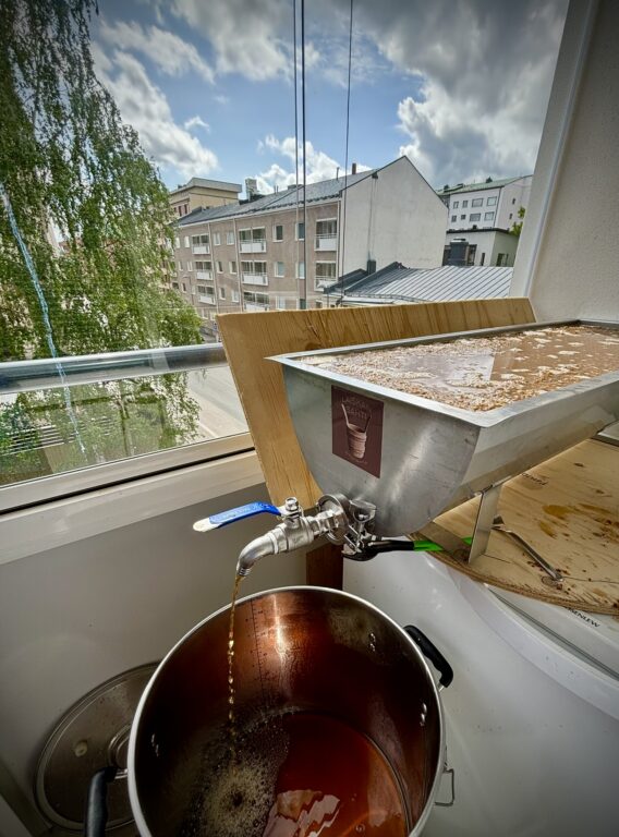 Kuva kerrostaloasunnon parvekkeelta. Jääkaapin päällä kaksi puuvaneria, joiden päällä metallinen kuurna, jossa mäskiä ja vettä. Ruskeaa nestettä valuu kuurnan hanasta alla olevaan isoon metalliastiaan. Taustalla näkyy iso puu, kerrostaloja ja pilvinen taivas.