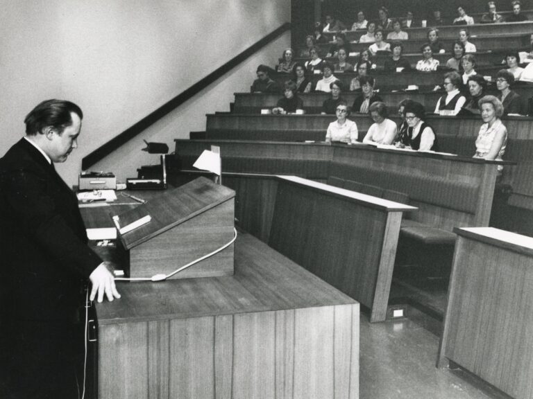 Vasemmalla mies seisoo luentosalin edessä puhujatelineen luona. Taustalla näkyy luentosalissa istuvia ihmisiä.